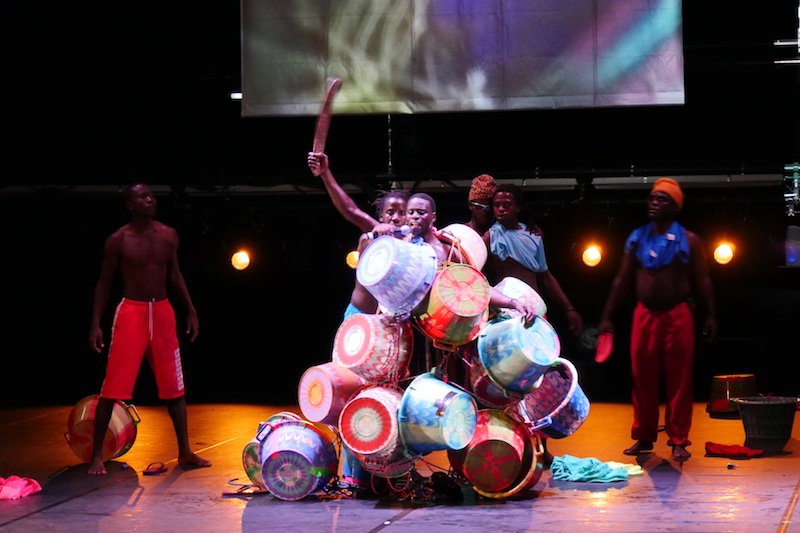Colorful baskets are arranged in a shoulder high mound as male performers interact behind it